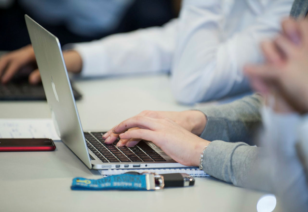 Person tippt auf Laptop-Tastatur während eines Meetings, Schreibblock und USB-Stick liegen auf Tisch.